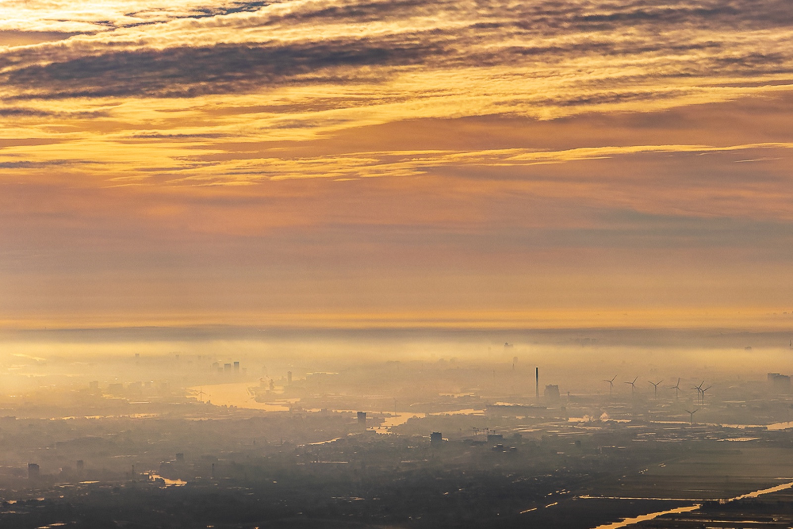Vista aérea de una ciudad tendida bajo una capa de niebla al amanecer de otoño; el cielo, estratificado en nubes doradas, ocupa la mitad superior, y abajo un estuario devuelve la luz entre grúas, chimeneas y aerogeneradores.